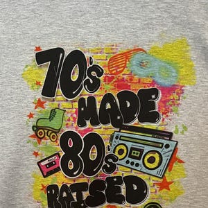 May include: Grey t-shirt featuring a graphic print of a brick wall with the text "70's made 80's raised" in black. The graphic also includes a roller skate, a cassette tape, and a boombox.