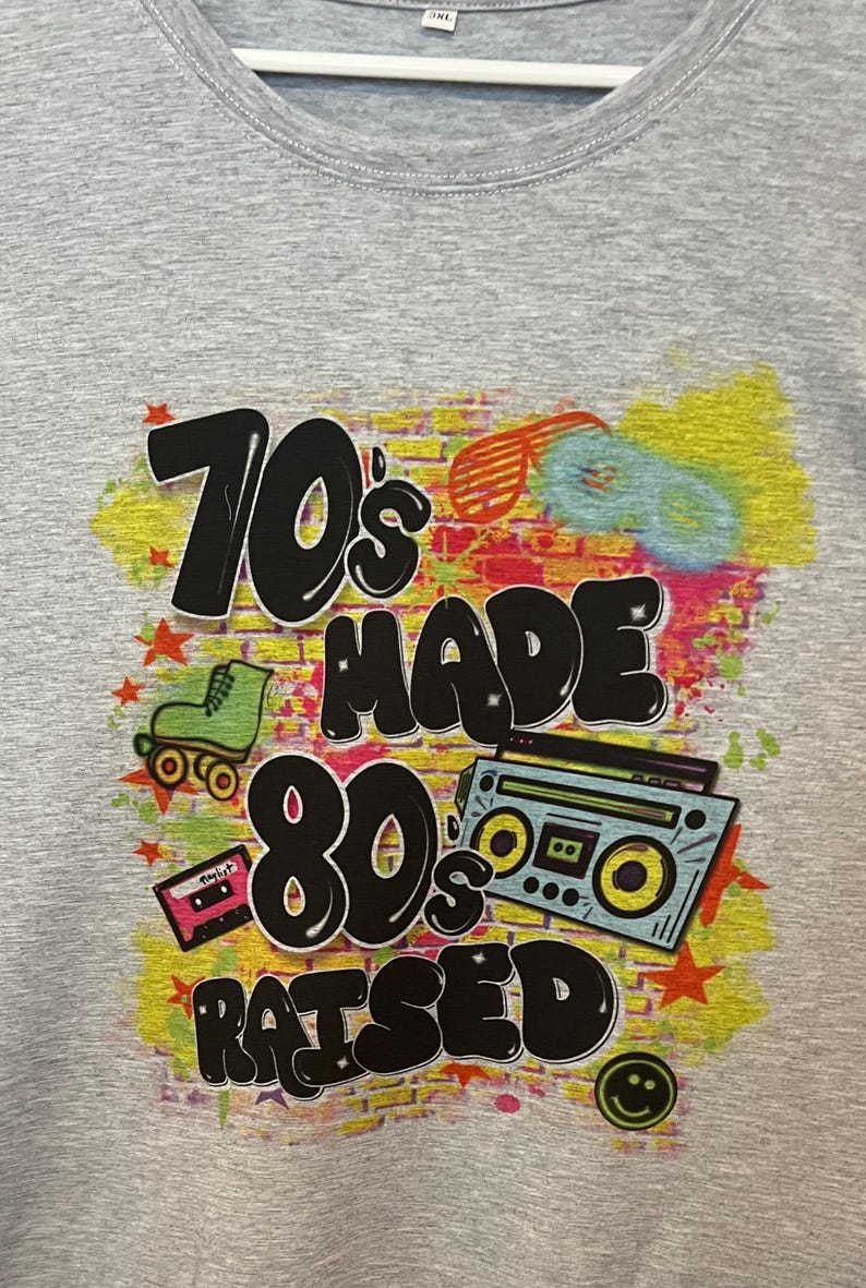 May include: Grey t-shirt featuring a graphic print of a brick wall with the text "70's made 80's raised" in black. The graphic also includes a roller skate, a cassette tape, and a boombox.