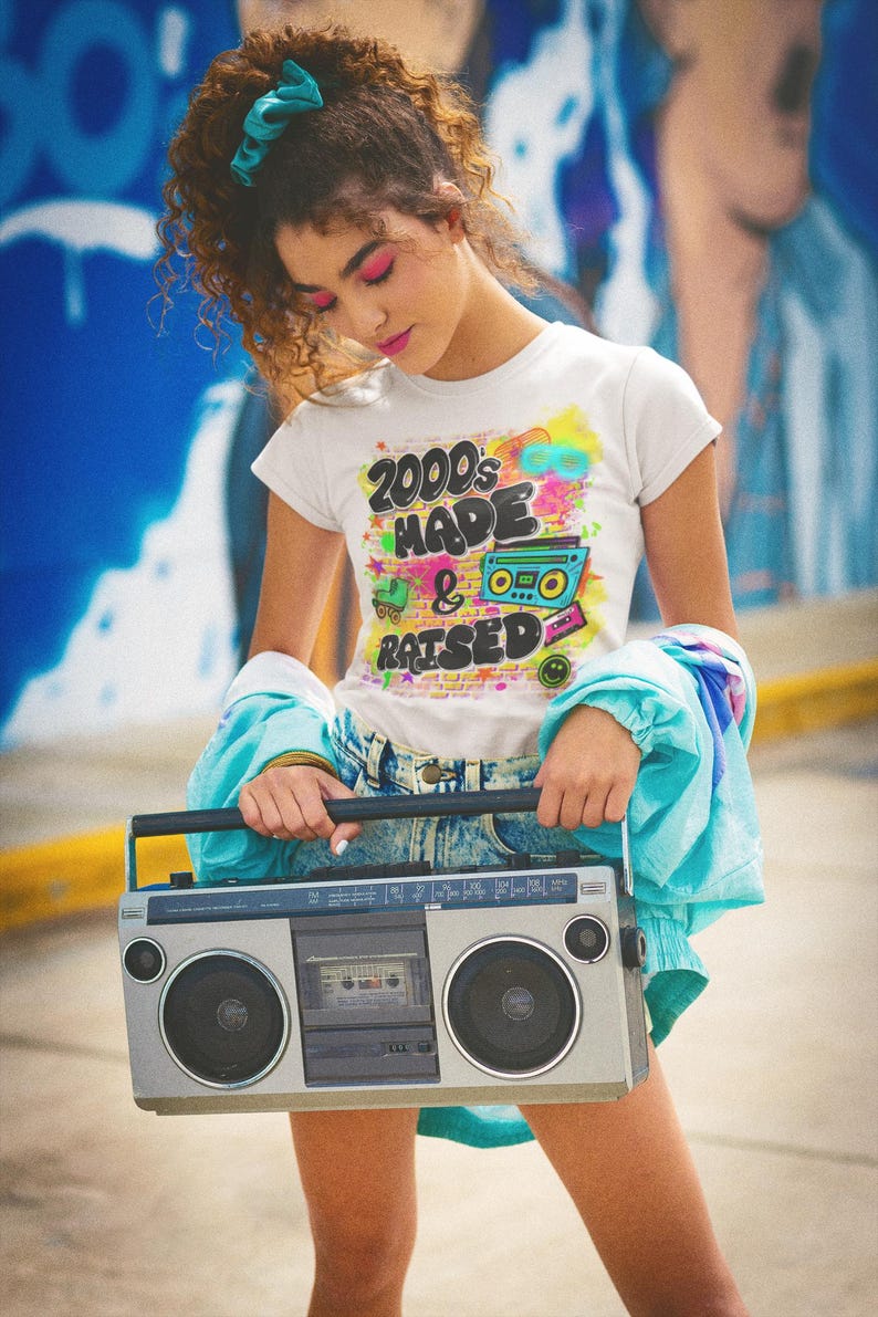 May include: A young person wearing a white t-shirt with the text "2000's Made & Raised" in colourful graffiti style. The person is holding a silver boombox with a cassette tape player.