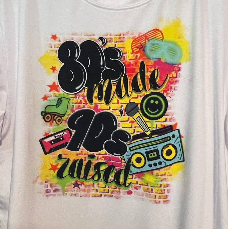 May include: A white t-shirt featuring a graphic design of a brick wall with the text "80's made 90's raised" in black and white. The graphic also includes a cassette tape, a microphone, roller skates, and a boombox.