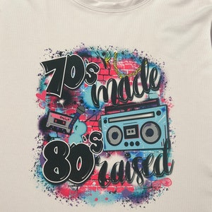 May include: A white t-shirt with a graphic design featuring a brick wall with a cassette tape, microphone, and a boombox. The text on the shirt reads "70's made 80's raised".