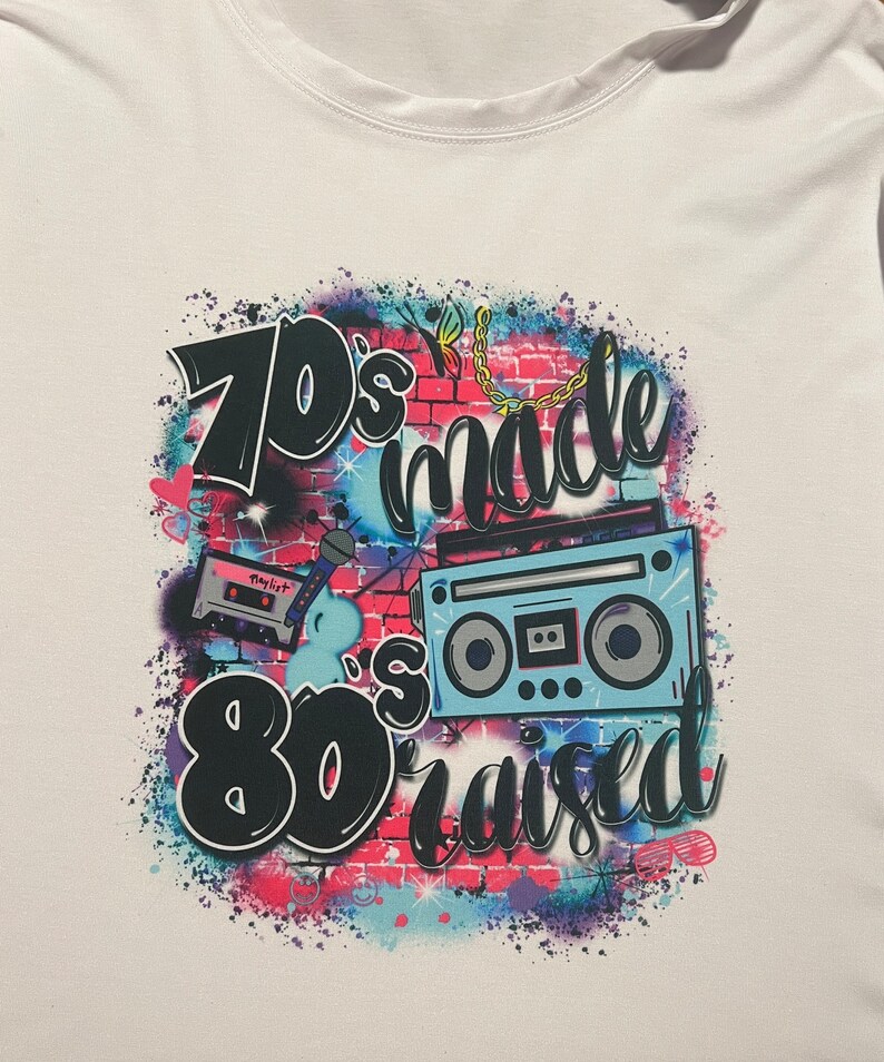 May include: A white t-shirt with a graphic design featuring a brick wall with a cassette tape, microphone, and a boombox. The text on the shirt reads "70's made 80's raised".