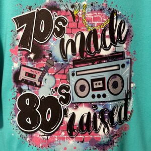 May include: A turquoise t-shirt with a graphic design featuring a brick wall, a cassette tape, a boombox, and the text "70's made 80's raised".