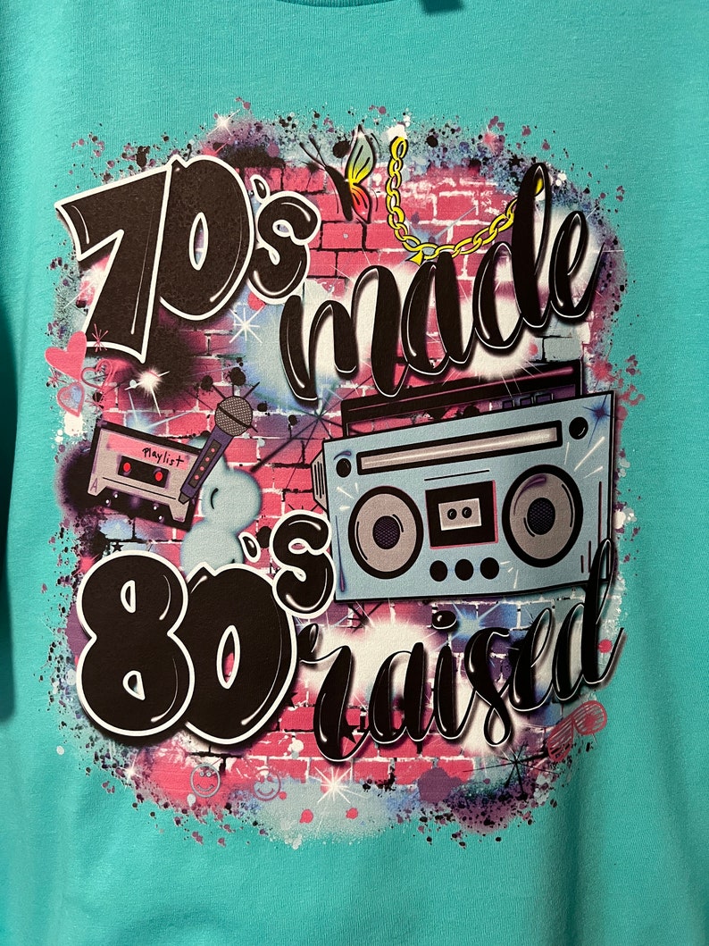 May include: A turquoise t-shirt with a graphic design featuring a brick wall, a cassette tape, a boombox, and the text "70's made 80's raised".