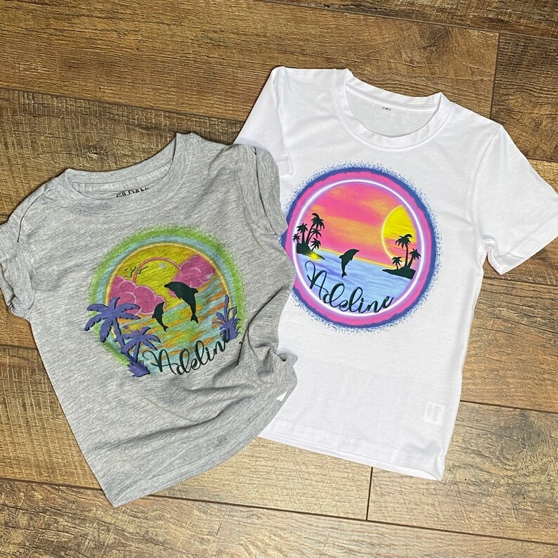 Pu&ograve; includere: Due magliette con un design di tramonto e delfini. La maglietta grigia ha il nome "Addie" stampato su di essa. La maglietta bianca ha il nome "Addie" stampato su di essa.