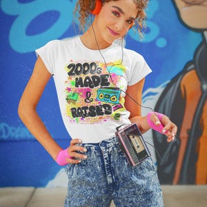 May include: A woman wearing a white t-shirt with the text "2000's Made & Raised" in colourful graffiti style. She is wearing blue acid wash jeans and holding a pink cassette player.