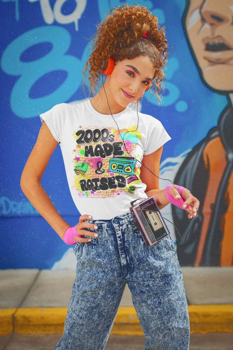 May include: A woman wearing a white t-shirt with the text "2000's Made & Raised" in colourful graffiti style. She is wearing blue acid wash jeans and holding a pink cassette player.