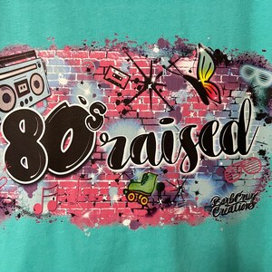 May include: A turquoise t-shirt with a graphic design of a brick wall with 80's themed imagery, including a boombox, roller skates, a butterfly, and the text "80's raised".