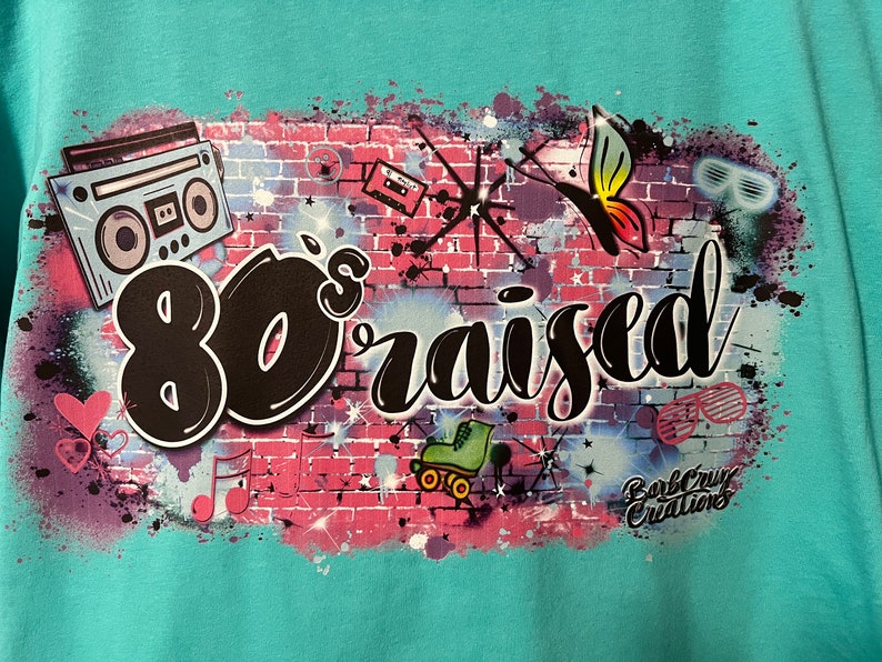 May include: A turquoise t-shirt with a graphic design of a brick wall with 80's themed imagery, including a boombox, roller skates, a butterfly, and the text "80's raised".