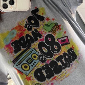 May include: A grey t-shirt with a colourful graphic print that says "Raised 80's Made 70's". The graphic includes a cassette tape, roller skates, and a boombox.