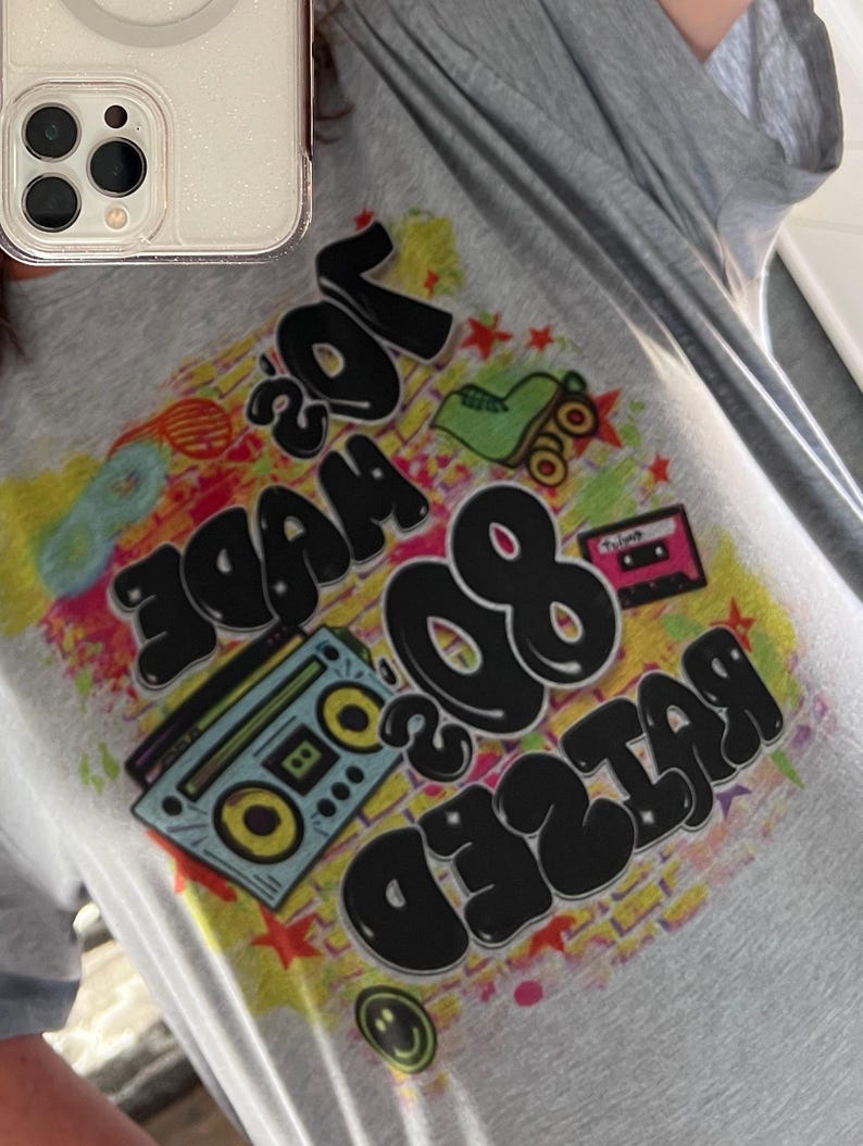 May include: A grey t-shirt with a colourful graphic print that says "Raised 80's Made 70's". The graphic includes a cassette tape, roller skates, and a boombox.