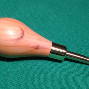 Könnte beinhalten: Ein Häkelhaken aus Holz mit einem hellbraunen Holzgriff und einem silbernen Metallhaken. Der Haken liegt auf einer grünen Oberfläche.