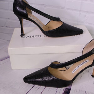 Puede incluir: Un par de zapatos de sal&oacute;n negros de cuero con punta puntiaguda y tac&oacute;n de aguja. Los zapatos tienen un dise&ntilde;o de corte en el empeine y est&aacute;n sobre una caja blanca con el logotipo de Manolo Blahnik.