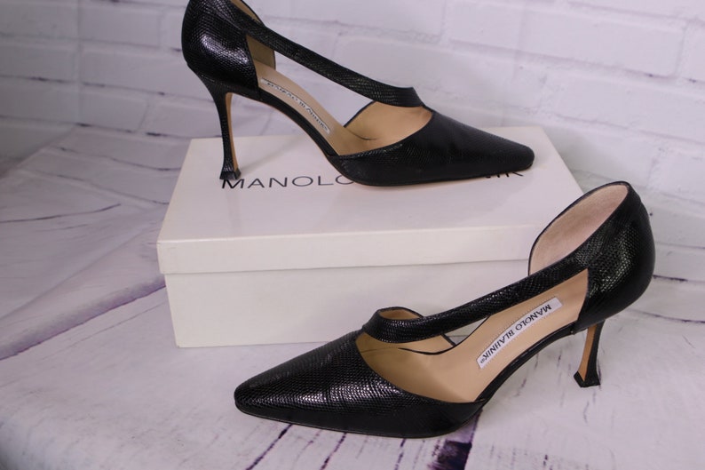 Puede incluir: Un par de zapatos de sal&oacute;n negros de cuero con punta puntiaguda y tac&oacute;n de aguja. Los zapatos tienen un dise&ntilde;o de corte en el empeine y est&aacute;n sobre una caja blanca con el logotipo de Manolo Blahnik.