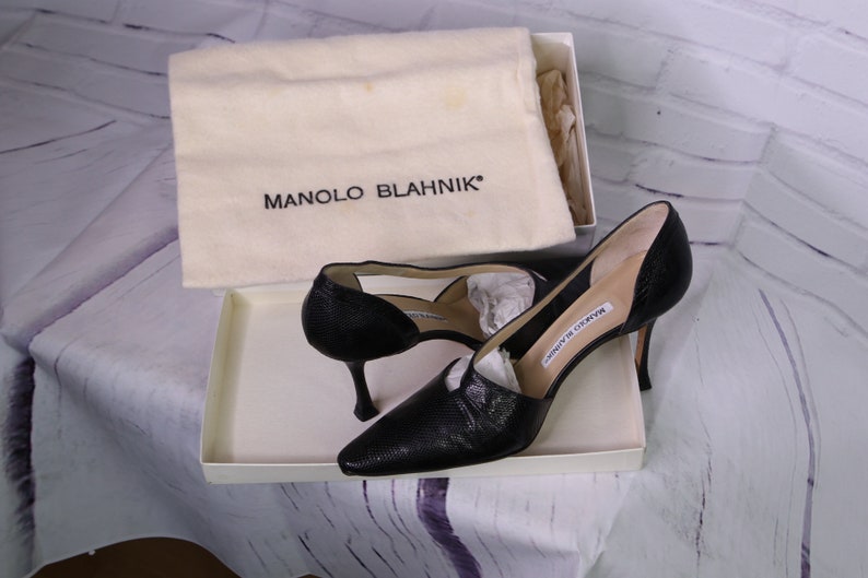 Puede incluir: Un par de zapatos de tac&oacute;n alto de cuero negro con punta puntiaguda y dise&ntilde;o abierto. Los zapatos est&aacute;n en una caja blanca con una bolsa de polvo que dice "MANOLO BLAHNIK".