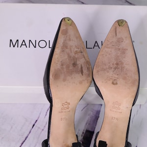 Puede incluir: Un par de zapatos de tac&oacute;n de cuero negro con punta puntiaguda. Los zapatos tienen una peque&ntilde;a correa alrededor del tobillo y est&aacute;n etiquetados "Manolo Blahnik" en las suelas. La talla es 37 1/2.