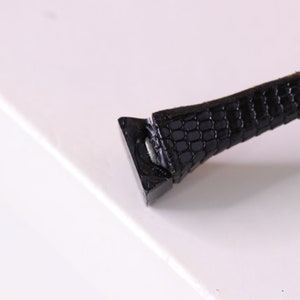 Puede incluir: Primer plano de una correa de cuero negra con un patr&oacute;n texturizado. La correa est&aacute; unida a una hebilla de metal.