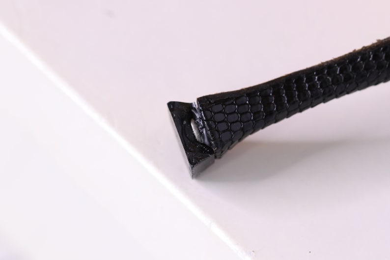 Puede incluir: Primer plano de una correa de cuero negra con un patr&oacute;n texturizado. La correa est&aacute; unida a una hebilla de metal.