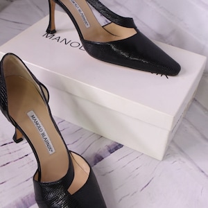 Puede incluir: Un par de zapatos de sal&oacute;n de cuero negro con punta puntiaguda y tac&oacute;n de aguja. Los zapatos tienen un dise&ntilde;o recortado en el empeine y est&aacute;n etiquetados como "Manolo Blahnik".