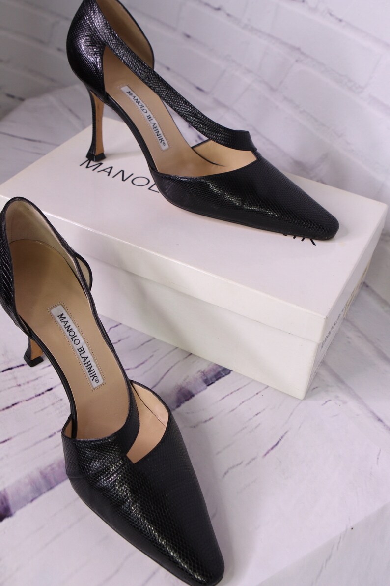 Puede incluir: Un par de zapatos de sal&oacute;n de cuero negro con punta puntiaguda y tac&oacute;n de aguja. Los zapatos tienen un dise&ntilde;o recortado en el empeine y est&aacute;n etiquetados como "Manolo Blahnik".