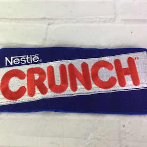 May include: A blue and white embroidered patch with the words "Nestle Crunch" in red letters.