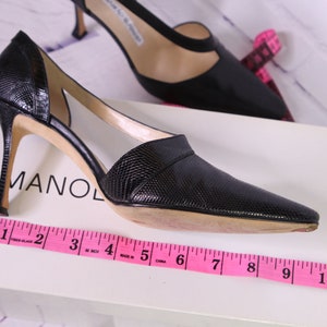 Puede incluir: Un par de zapatos de tac&oacute;n de cuero negro con un dise&ntilde;o recortado. Los zapatos est&aacute;n sobre una caja blanca con la palabra "MANOL" visible. Una cinta m&eacute;trica rosa est&aacute; colocada sobre la caja, mostrando que la altura del tac&oacute;n es de 7,6 cm.