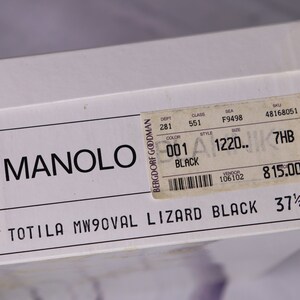 Puede incluir: Una caja de zapatos blanca con texto negro que dice "MANOLO BERGDORF GOODMAN C TOTILA MW9OVAL LIZARD BLACK 37 1/2". Una etiqueta blanca m&aacute;s peque&ntilde;a con texto negro est&aacute; pegada a la caja. La etiqueta dice "DEPT 281 CLASS 551 SEA F9498 SKU 48168051 COLOR 001 STYLE 1220.. SIZE 7HB BLACK VENDOR 106102 815.00"