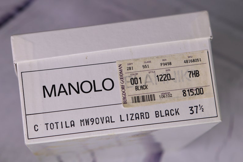 Puede incluir: Una caja de zapatos blanca con texto negro que dice "MANOLO BERGDORF GOODMAN C TOTILA MW9OVAL LIZARD BLACK 37 1/2". Una etiqueta blanca m&aacute;s peque&ntilde;a con texto negro est&aacute; pegada a la caja. La etiqueta dice "DEPT 281 CLASS 551 SEA F9498 SKU 48168051 COLOR 001 STYLE 1220.. SIZE 7HB BLACK VENDOR 106102 815.00"