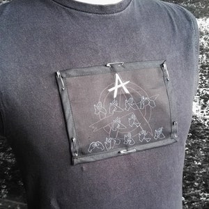 May include: A black t-shirt with a black patch featuring a white anarchy symbol and several white hand gestures. The patch is attached to the shirt with safety pins.
