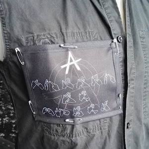 May include: Black sleeveless shirt with a black patch featuring a white anarchy symbol and white hand signs. The patch is attached to the shirt with safety pins.