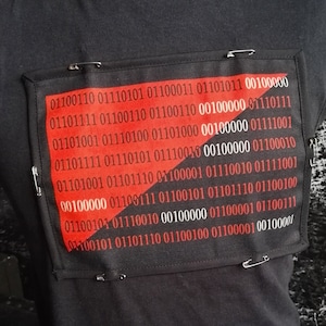 Peut inclure: T-shirt noir avec un patch rouge et noir représentant du code binaire. Le patch est fixé avec des épingles de sûreté. Le design du t-shirt comprend le code binaire : 01100110 01110101 01100011 01101011 00100000 01101111 01100110 01100110 00100000 01110111.