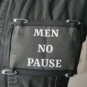 May include: A black fabric patch with white text that reads "MEN NO PAUSE" is pinned to a black jacket with safety pins. The patch is surrounded by four safety pins.