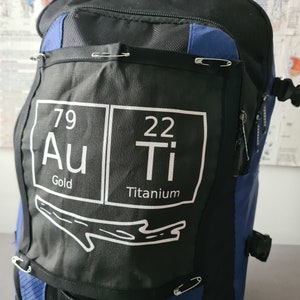 Peut inclure: Sac à dos noir et bleu avec un patch blanc représentant les éléments du tableau périodique pour l'or (Au) et le titane (Ti). Le patch comprend également les mots "Or" et "Titane" sous les éléments.