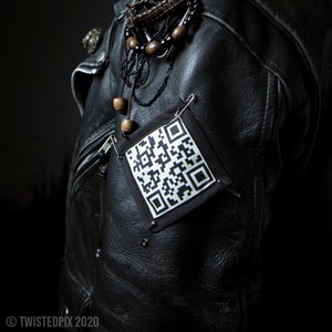 May include: A black leather jacket with a black and white QR code patch attached to the left sleeve. The jacket has a beaded necklace with wooden beads and a silver chain.