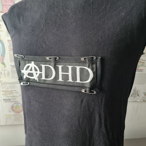 May include: Black sleeveless shirt with a black and white patch that says "ADHD" with an anarchy symbol. The patch is attached to the shirt with safety pins.
