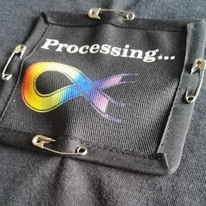 May include: A black fabric patch with a rainbow infinity symbol and the text "Processing..." attached to a black shirt with safety pins.