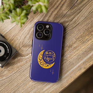 May include: A dark blue phone case featuring a gold crescent moon design with an intricate symbol inside. The case has a glossy finish and includes gold star accents. The phone case is resting on a wooden surface.