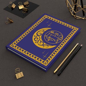 May include: A dark blue journal with a gold crescent moon and sigil design on the cover. The cover is framed with a gold border. Gold pencils and binder clips are next to the journal.