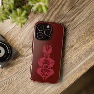 May include: A claret-coloured phone case with a decorative red design featuring abstract shapes and symbols. The case is on a wooden surface, with a camera and plant in the background.