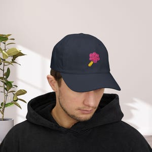 May include: A navy blue baseball cap featuring an embroidered pink and yellow popsicle design. The cap has a curved peak and a classic six-panel design. The design shows a pink popsicle with a yellow stick.