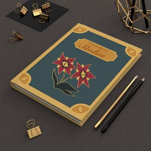 May include: A rectangular hardcover journal with a dark teal cover, gold borders, and four gold eye-shaped corner accents. The center features two red flowers with yellow eye-shaped centers and the text Property of Stolas in gold script.