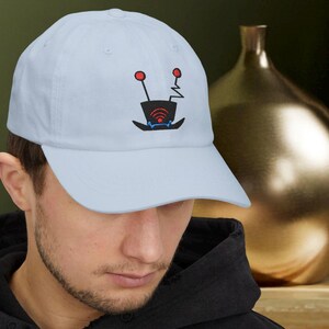 Peut inclure: Casquette de baseball bleu clair avec un motif brodé. Le motif représente un chapeau haut de forme noir avec des cercles rouges en forme d'antenne et un symbole Wi-Fi. Le chapeau est de style classique et réglable.