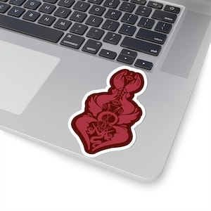 May include: A maroon sticker with a detailed, stylized design featuring a skull, geometric shapes, and ornate elements. The sticker is placed on a silver laptop, showcasing its shape and colour.