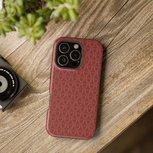 Peut inclure: Coque de téléphone rouge avec un motif répété de cœurs. Le boîtier a un bord noir autour des lentilles de la caméra et un bord noir assorti. La coque est posée sur une surface en bois.