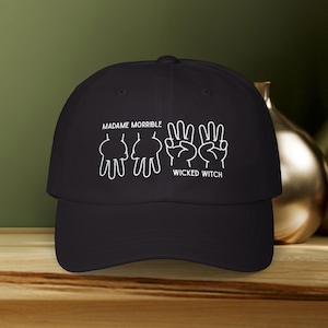 May include: Black baseball cap with white text and a hand gesture design. The text reads "MADAME MORRIBLE" above a series of hand gestures, and "WICKED WITCH" below. The cap is on a wooden surface.