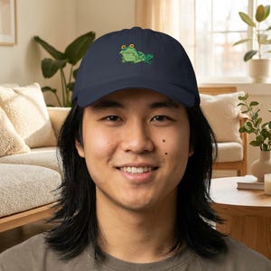 May include: Navy blue baseball cap featuring a green frog graphic with yellow eyes. The frog is embroidered on the front of the cap. The cap is made of a soft, durable material and has a curved brim.