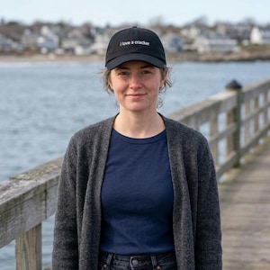 Op de afbeelding: Zwarte baseballcap met de tekst "I love a cracker" in het wit. De cap wordt gedragen door een persoon. De persoon draagt een marineblauw t-shirt en een grijze cardigan. De achtergrond toont een water en een houten pier.