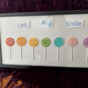 May include: A rectangular black frame containing seven colorful swirl lollipops arranged in a horizontal row. Above the lollipops, three white rectangular labels feature the blue printed text Lolli, Pop, and Smile. A small signature is in the corner.