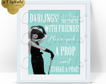 Op de afbeelding: Een ingelijst bord met een lichtblauwe achtergrond en een zwart-witillustratie van een persoon in een zwarte jurk, hoed en handschoenen met rode lippen. De tekst luidt: DARLINGS! IT'S TIME FOR PHOTOS WITH FRIENDS! PLEASE GRAB A PROP AND STRIKE A POSE!