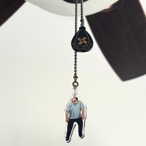 May include: A ceiling fan pull chain with a black and bronze Harbor Breeze logo and a clear acrylic charm featuring a cartoon image of a man in a blue shirt and black pants.